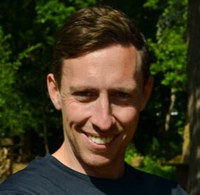 Man with brown hair smiling at camera outdoors with green foliage background